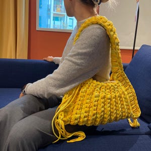 May include: A bright yellow, handmade crochet bag with a thick strap. The bag is a vibrant yellow color and appears to be made from a soft, textured yarn. The strap is also yellow and is designed to be worn over the shoulder.