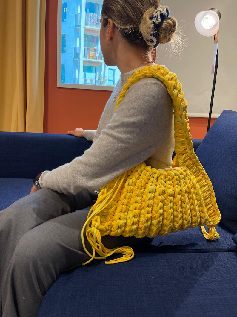 May include: A bright yellow, handmade crochet bag with a thick strap. The bag is a vibrant yellow color and appears to be made from a soft, textured yarn. The strap is also yellow and is designed to be worn over the shoulder.