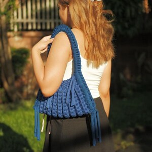 May include: A blue macrame shoulder bag with a braided strap and fringe detail. The bag is carried over the shoulder. The person is wearing a white tank top and black skirt. A flower is in the person's hair.
