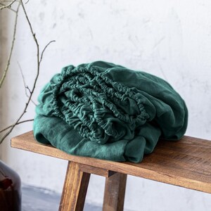 Könnte beinhalten: Ein gefaltetes, dunkelgrünes Spannbettlaken liegt auf einer rustikalen Holzbank. Links steht eine braune Glasvase mit kahlen Ästen. Die Bank steht vor einer weißen Wand und einem grauen Boden. Der Text "Linen HUE" ist sichtbar.