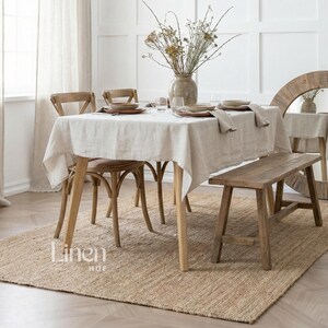 Op de afbeelding: Een rechthoekige eettafel bedekt met een lichtbeige linnen tafelkleed, voorzien van tafelinstellingen met keramische borden en servetten. De tafel is omringd door houten stoelen en een houten bank, met een vaas met droogbloemen als middelpunt.