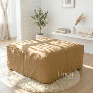 Könnte beinhalten: Ein großer, quadratischer Hocker in einem warmen Beigeton steht auf einem flauschigen, cremefarbenen Teppich. Der Hocker besteht aus einem strukturierten Stoff und steht auf einem hellen Holzboden. Die Worte "Linen HUE" sind unten rechts zu sehen.