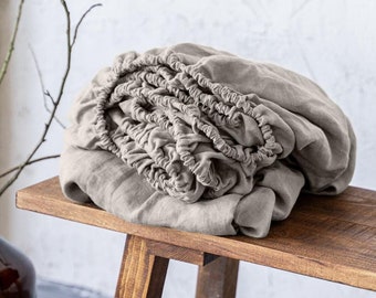 Puede incluir: Una sábana bajera de lino beige está doblada y colocada sobre un banco de madera. El banco tiene un acabado rústico y desgastado. El texto "Linen Hue" es visible en el banco.