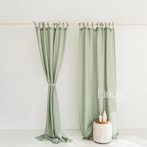 May include: Sage green linen curtains hanging from a wooden rod. One curtain is tied back with a white cord. The curtains are in front of a white wall. The Linen Hue logo is visible on the right curtain.