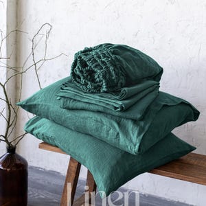 Könnte beinhalten: Ein Stapel dunkelgrüner Bettwäsche aus Leinen, bestehend aus einem Spannbettlaken, einem Bettlaken und Kissenbezügen, gefaltet und auf einer Holzbank präsentiert. Die Bettwäsche ist aus 100% Leinen gefertigt und weich, atmungsaktiv und strapazierfähig. Der Text "Linen Hue" ist auf der Bank sichtbar.