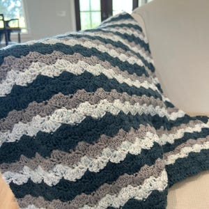 May include: A handmade crochet blanket in a shell stitch pattern. The blanket features alternating rows of dark teal, light gray, white, and medium gray. The blanket is draped over a light-colored armchair.