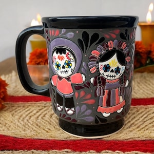 May include: A gray ceramic mug featuring a black handle and base, decorated with two raised figures painted in the style of Day of the Dead calaveras. The figures are surrounded by colorful floral and leaf motifs in shades of red, purple, and blue.
