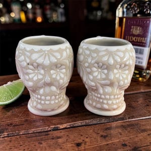 May include: Two ceramic shot glasses shaped like skulls, featuring a light beige finish with raised white floral and leaf patterns. The design includes detailed teeth and eye sockets, with a smooth, glossy texture covering the entire surface.