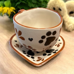 May include: A ceramic teacup and saucer set with a paw print design. The cup and saucer are off-white with brown accents and paw prints. The saucer is heart-shaped. The cup has a small handle.