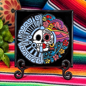 May include: A square black ceramic tile featuring a circular design of a skull split into two halves. The left side displays blue geometric patterns, while the right side features colorful floral motifs in red, yellow, and purple. The tile rests on a black metal stand.
