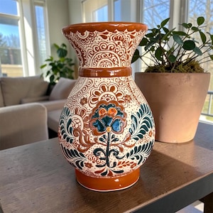 May include: A ceramic vase with a flared rim and a rounded body, featuring a cream-colored base decorated with intricate brown and dark green floral and paisley patterns. The vase has a solid brown band around the neck and a solid brown base.
