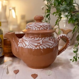 May include: A brown ceramic teapot with a lid, adorned with white floral patterns. The teapot has a curved spout and handle. A jar of Nescafé coffee is visible to the left. The scene is set on a white surface with decorative elements.