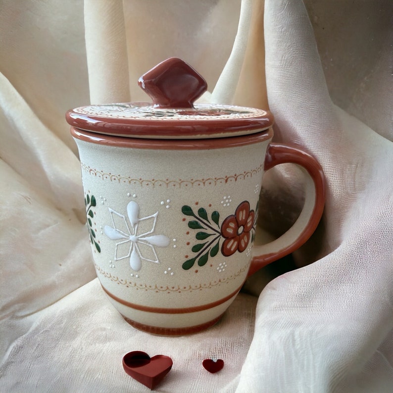 May include: A beige ceramic mug with a lid, decorated with floral and geometric patterns in white, green, and brown. The mug has a handle and sits on a cream-coloured fabric. Two small red paper hearts are in the foreground.