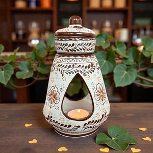 May include: A decorative ceramic oil warmer with a lid, featuring a cream base and brown floral patterns. The warmer has a large opening and a lit candle inside, creating a warm ambiance. The design includes a brown top and base.