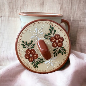 May include: A ceramic mug with a matching lid. The lid is beige with a brown rim and handle, decorated with red flowers, white accents, and green leaves. The mug is light green with a brown rim.