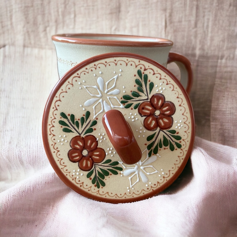 May include: A ceramic mug with a matching lid. The lid is beige with a brown rim and handle, decorated with red flowers, white accents, and green leaves. The mug is light green with a brown rim.