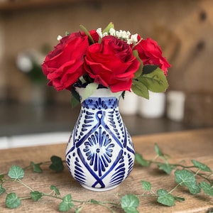 May include: A white and blue patterned vase holds a bouquet of vibrant red roses. The vase has a detailed design and sits on a wooden surface. Green ivy-like vines are draped around the vase, adding a natural touch. The background is blurred.