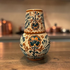 May include: A ceramic carafe and cup set featuring a cream-colored base with intricate hand-painted floral and foliate patterns in deep teal and burnt orange. The cup sits inverted atop the rounded carafe, creating a cohesive decorative vessel.