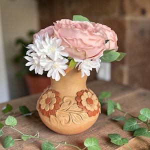 May include: A terracotta vase with a floral design, holding a bouquet of pink roses and white daisies. The vase has a detailed relief pattern of flowers and leaves. Green vines are draped around the vase on a wooden table.
