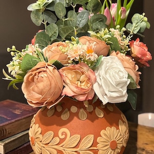May include: A terracotta-colored vase with a floral design holds a bouquet of artificial flowers. The bouquet includes peach-colored peonies, white roses, and greenery. The vase has a detailed embossed floral pattern.