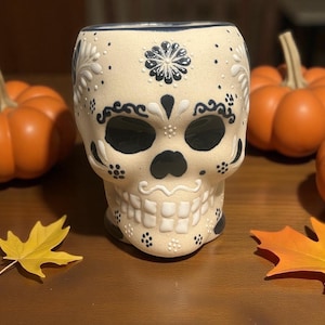 May include: A cream-colored ceramic mug shaped like a human skull, featuring black and white painted floral and geometric patterns. The skull has deep black eye sockets, a black nose cavity, and raised white teeth, with a smooth matte finish.