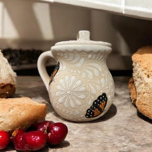May include: A cream-colored ceramic jar with a lid and handle, featuring embossed white floral and leaf patterns. Two monarch butterflies with orange, black, and white wings are painted on the rounded body of the jar, which has a glossy finish.