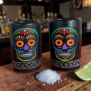 May include: Two black ceramic shot glasses featuring a colorful sugar skull design with red, orange, purple, and blue accents. Each glass has a white swirling pattern around the base, accompanied by a small pile of salt and a lime wedge.