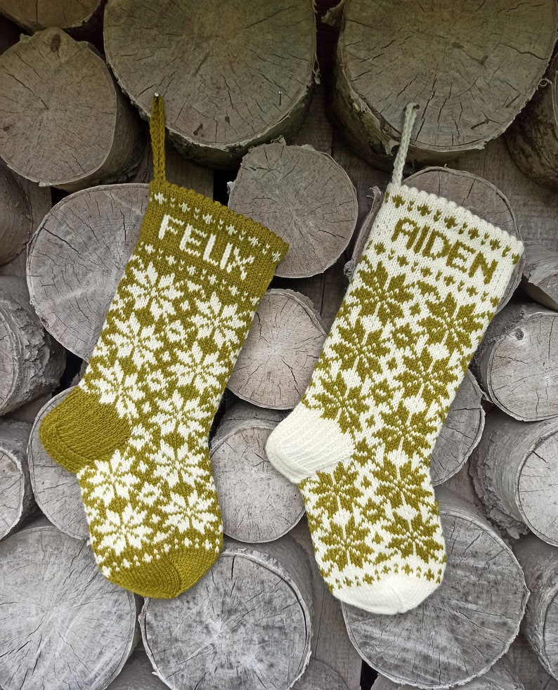 May include: Two knit Christmas stockings featuring a fair isle snowflake pattern in olive green and cream. The left stocking is primarily olive green with the name FELIX in cream, while the right stocking is primarily cream with the name AIDEN in olive green.