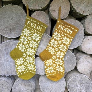 May include: Two mustard yellow and white knit Christmas stockings featuring a repeating snowflake pattern. The left stocking has the name FELIX knitted in white at the top, and the right stocking has the name ADAM knitted in white at the top.