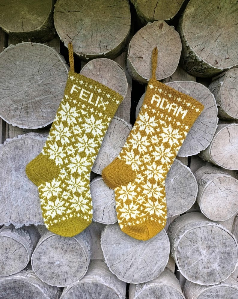 May include: Two mustard yellow and white knit Christmas stockings featuring a repeating snowflake pattern. The left stocking has the name FELIX knitted in white at the top, and the right stocking has the name ADAM knitted in white at the top.