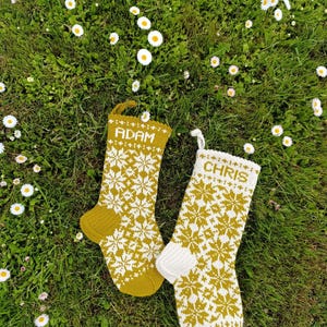 May include: Two knitted Christmas stockings featuring a repeating white snowflake pattern on a mustard yellow background. One stocking has the name ADAM in white text, while the other features the name CHRIS in white text, both with hanging loops.
