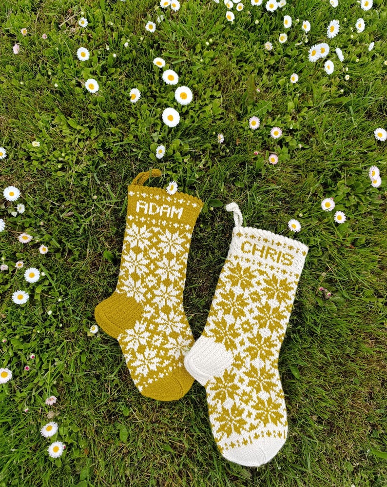 May include: Two knitted Christmas stockings featuring a repeating white snowflake pattern on a mustard yellow background. One stocking has the name ADAM in white text, while the other features the name CHRIS in white text, both with hanging loops.