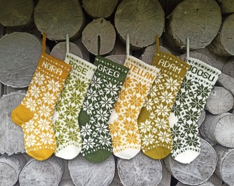 May include: Six knitted Christmas stockings featuring intricate white snowflake patterns on mustard yellow, olive green, and cream backgrounds. Each stocking is personalized with a name in white block letters: NORA, ROSE, GREG, STELLAN, ADAM, and JOOSU.