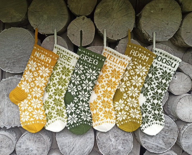 May include: Six knitted Christmas stockings featuring intricate white snowflake patterns on mustard yellow, olive green, and cream backgrounds. Each stocking is personalized with a name in white block letters: NORA, ROSE, GREG, STELLAN, ADAM, and JOOSU.