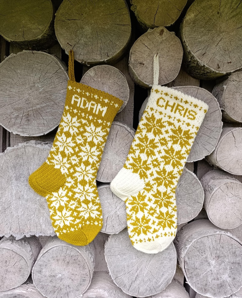 May include: Two knitted Christmas stockings featuring a repeating white and mustard yellow snowflake pattern. One stocking is primarily mustard yellow with the name ADAM in white, and the other is primarily white with the name CHRIS in mustard yellow.