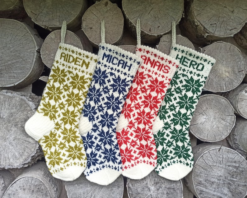 May include: Four knitted Christmas stockings featuring a white base with intricate snowflake patterns in olive green, navy blue, red, and dark green. Each stocking is personalized with the names AIDEN, MICAH, ANAIS, and HERO in block letters.