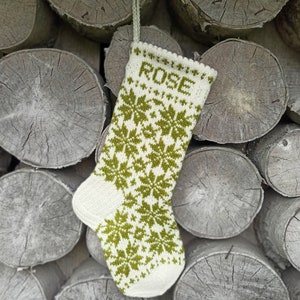 May include: A cream-colored knit Christmas stocking featuring a repeating olive green snowflake pattern and the name ROSE knitted in olive green letters at the top. The stocking includes a matching cream-colored knit hanging loop at the cuff.