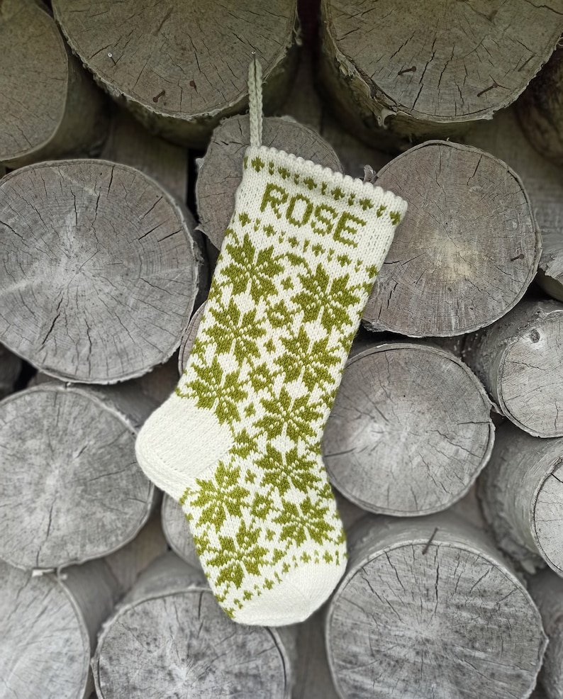 May include: A cream-colored knit Christmas stocking featuring a repeating olive green snowflake pattern and the name ROSE knitted in olive green letters at the top. The stocking includes a matching cream-colored knit hanging loop at the cuff.
