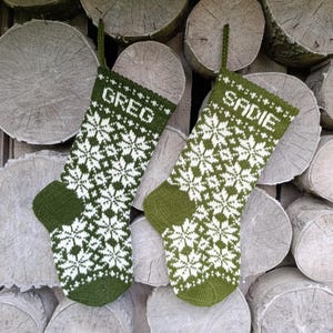 May include: Two olive green and white knit stockings featuring a repeating snowflake pattern. The left stocking has the name GREG knitted into the cuff, and the right stocking has the name SADIE knitted into the cuff. Both have a hanging loop.