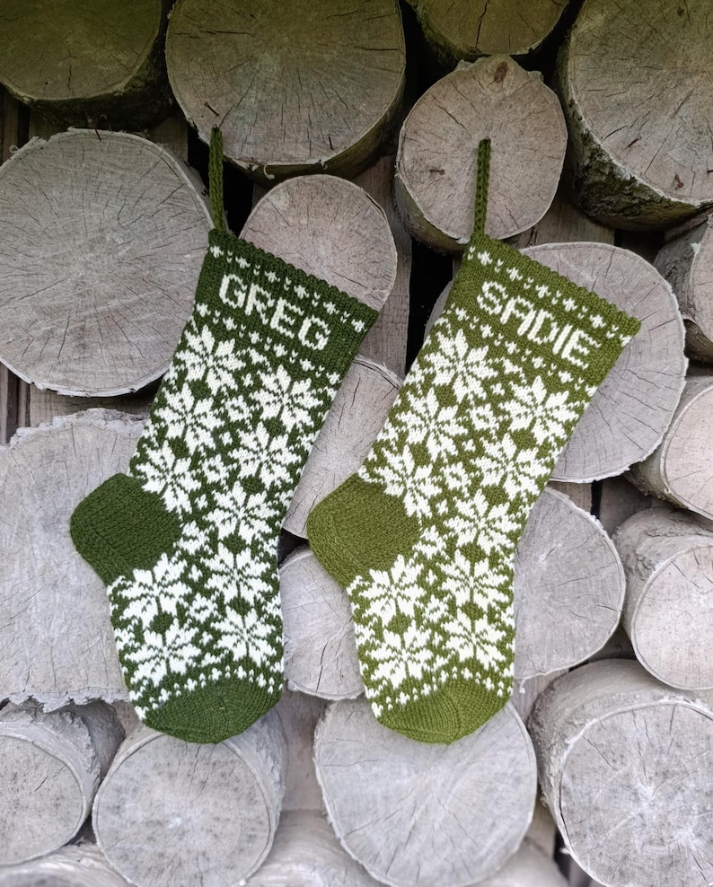 May include: Two olive green and white knit stockings featuring a repeating snowflake pattern. The left stocking has the name GREG knitted into the cuff, and the right stocking has the name SADIE knitted into the cuff. Both have a hanging loop.