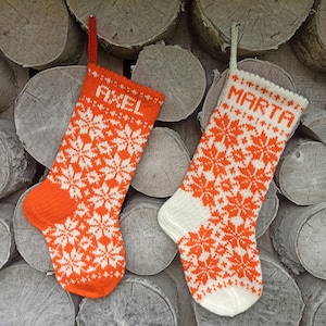 May include: Two orange and white knitted Christmas stockings with snowflake patterns. The stockings are personalized with the names "Axel" and "Marta".
