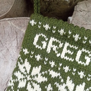 May include: A dark olive green knitted Christmas stocking featuring a white fair isle pattern and the name GREG knitted in white capital letters across the top section. The stocking includes a matching green yarn loop for hanging purposes.