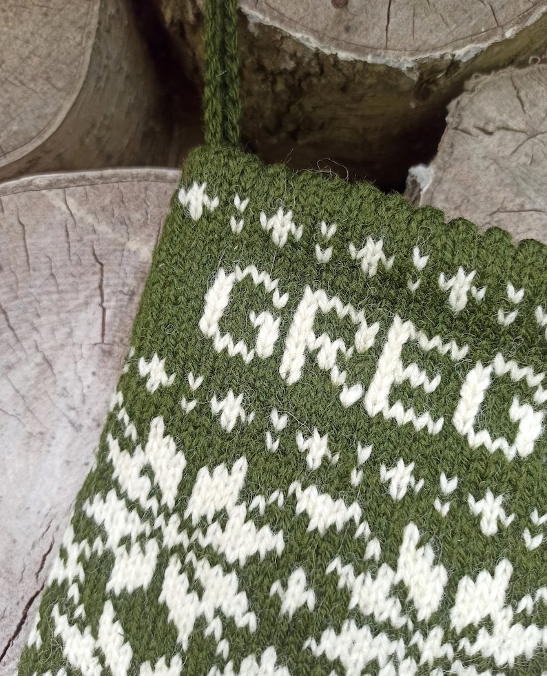 May include: A dark olive green knitted Christmas stocking featuring a white fair isle pattern and the name GREG knitted in white capital letters across the top section. The stocking includes a matching green yarn loop for hanging purposes.