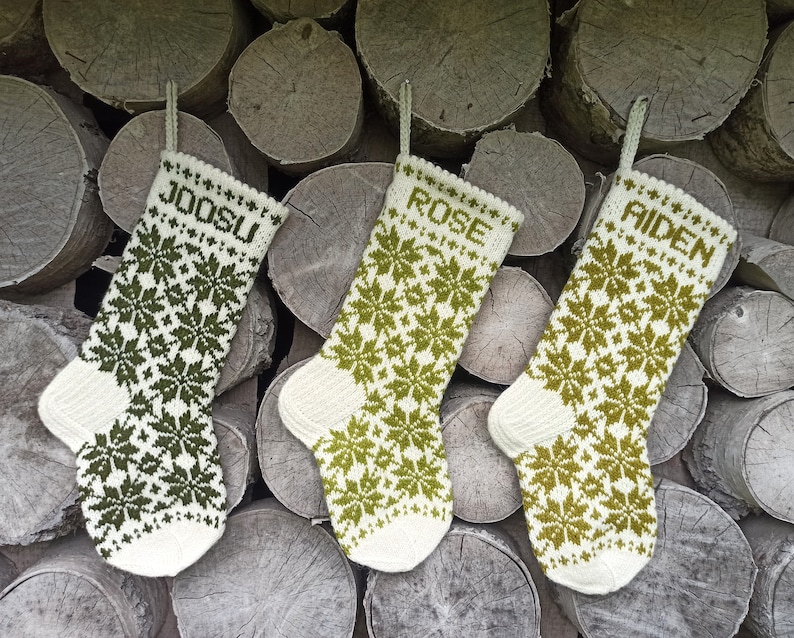 May include: Three knitted Christmas stockings featuring a white and green snowflake pattern. Each stocking has a hanging loop and is personalized with the names JOOSU, ROSE, and AIDEN in green block letters at the top of the cuff.
