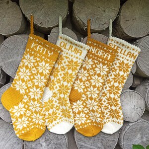 May include: Four knitted Christmas stockings featuring a repeating white and mustard yellow snowflake pattern. Each stocking is personalized with a name at the top: Katy, Olivia, Nora, and Stellan. The stockings have hanging loops and solid-colored toes.