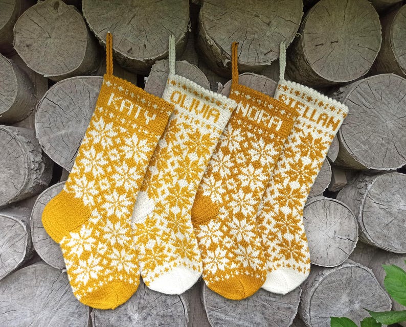 May include: Four knitted Christmas stockings featuring a repeating white and mustard yellow snowflake pattern. Each stocking is personalized with a name at the top: Katy, Olivia, Nora, and Stellan. The stockings have hanging loops and solid-colored toes.