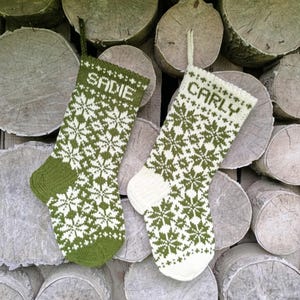 May include: Two knitted stockings featuring a repeating snowflake pattern. The left stocking is olive green with white details and the name SADIE. The right stocking is white with olive green details and the name CARLY. Both have hanging loops.