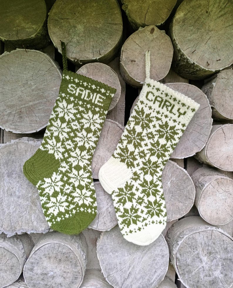 May include: Two knitted stockings featuring a repeating snowflake pattern. The left stocking is olive green with white details and the name SADIE. The right stocking is white with olive green details and the name CARLY. Both have hanging loops.