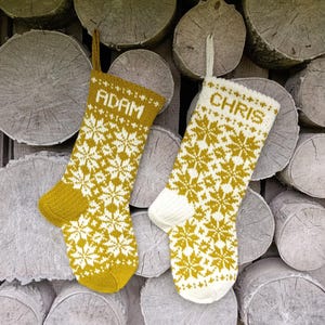 May include: Two knitted Christmas stockings featuring a repeating white and mustard yellow snowflake pattern. One stocking is primarily mustard yellow with the name ADAM in white, and the other is primarily white with the name CHRIS in mustard yellow.