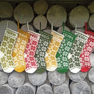 May include: Eight knitted Christmas stockings featuring a repeating snowflake pattern in various colors including olive green, mustard yellow, red, and white. Each stocking is personalized with a name at the top: Aiden, Nora, Nais, Greg, Stellan, Pearl, Joosu, and Nora.